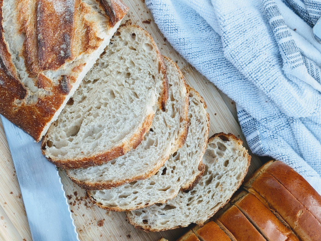 Sliced sourdough bread on a wooden board with a knife and a towel.