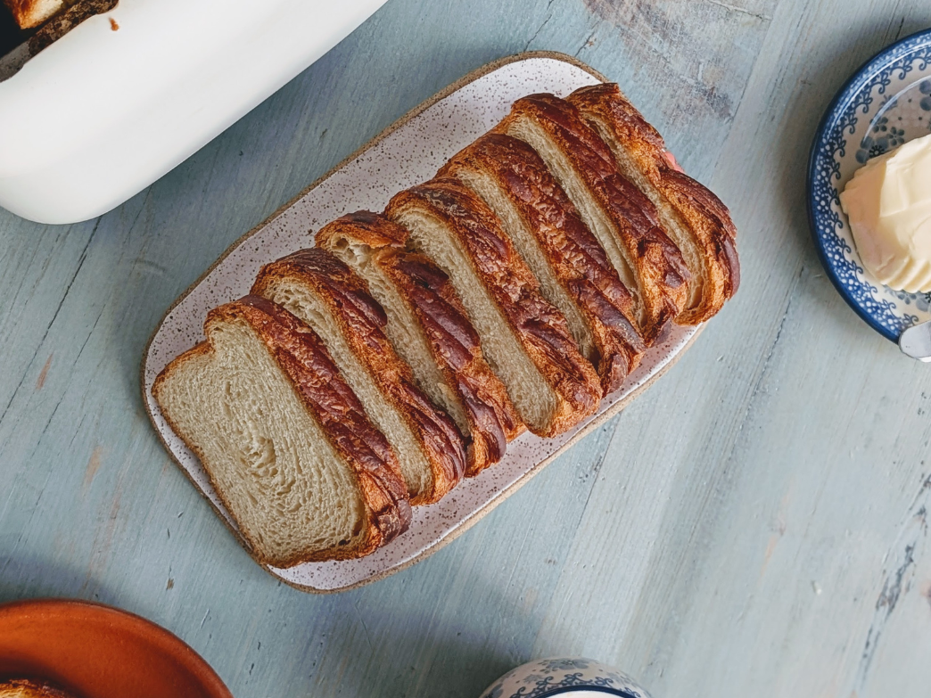 Sliced braided bread on a rectangular speckled plate on a blue wooden table with a dish of butter nearby
