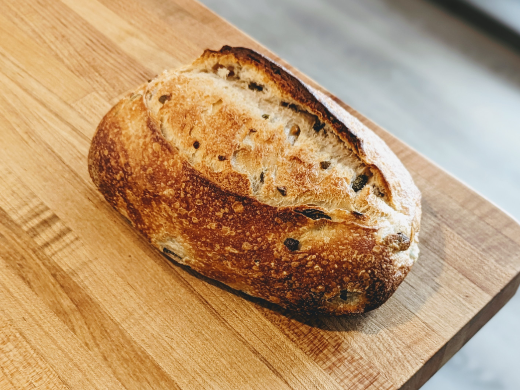 Angled top view of a whole crusty sourdough loaf with olive pieces and deep golden scoring on a wooden cutting board