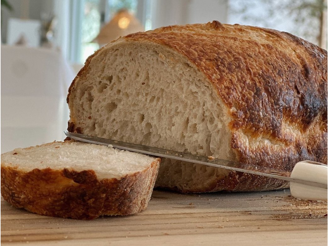 Crusty artisan loaf with one sliced piece and a serrated knife resting on a wooden cutting board.