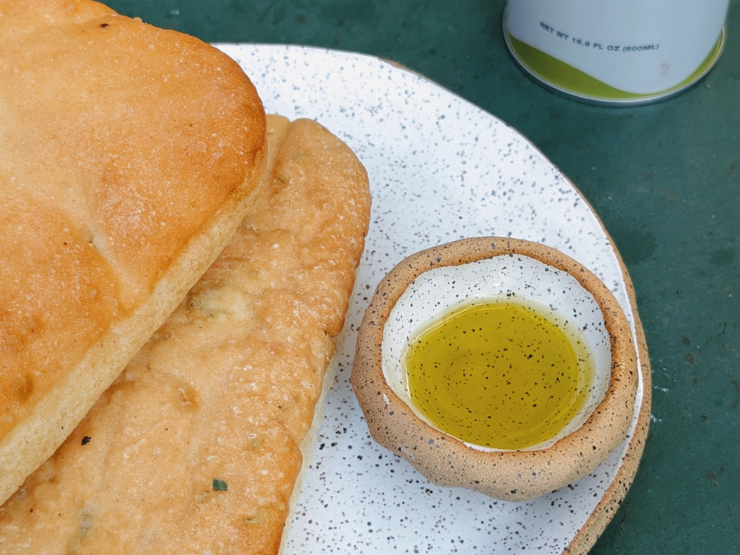 Small dish of olive oil with bread on a speckled plate; can text: NET WT 16.9 FL OZ (500ML)