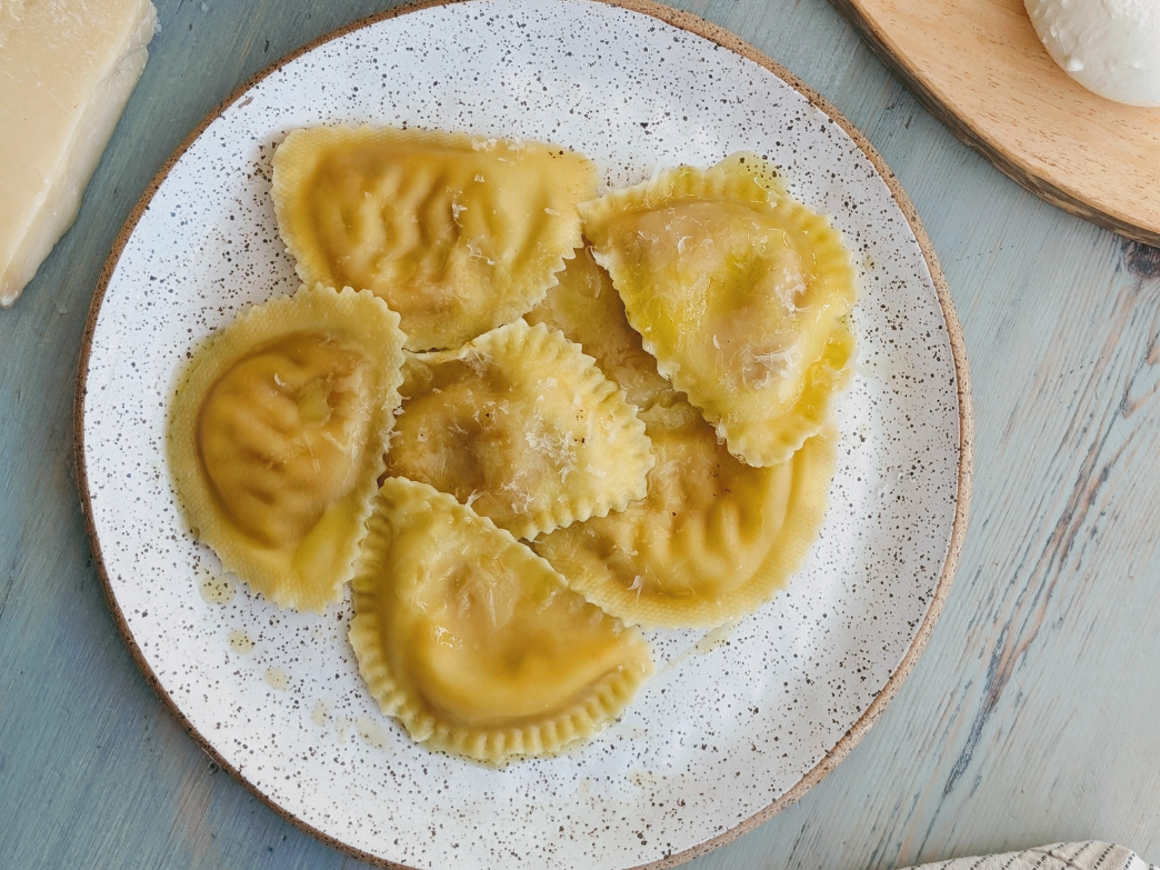 Six buttered ravioli with grated cheese on a white speckled plate over a blue wooden table