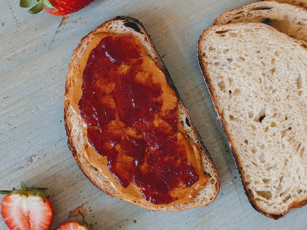 Slice of sourdough with peanut butter and strawberry jam spread, another plain slice and halved strawberries on wooden board