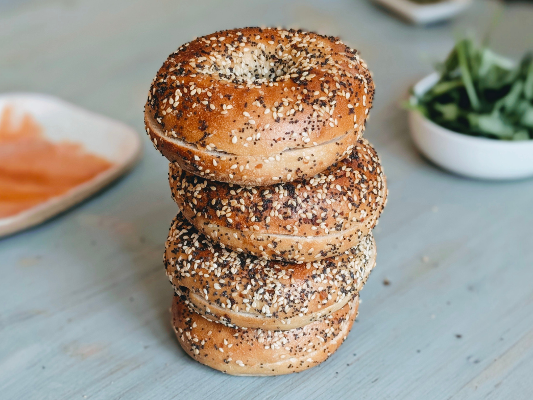 Stack of four everything bagels on a blue wooden table with smoked salmon and greens blurred in background