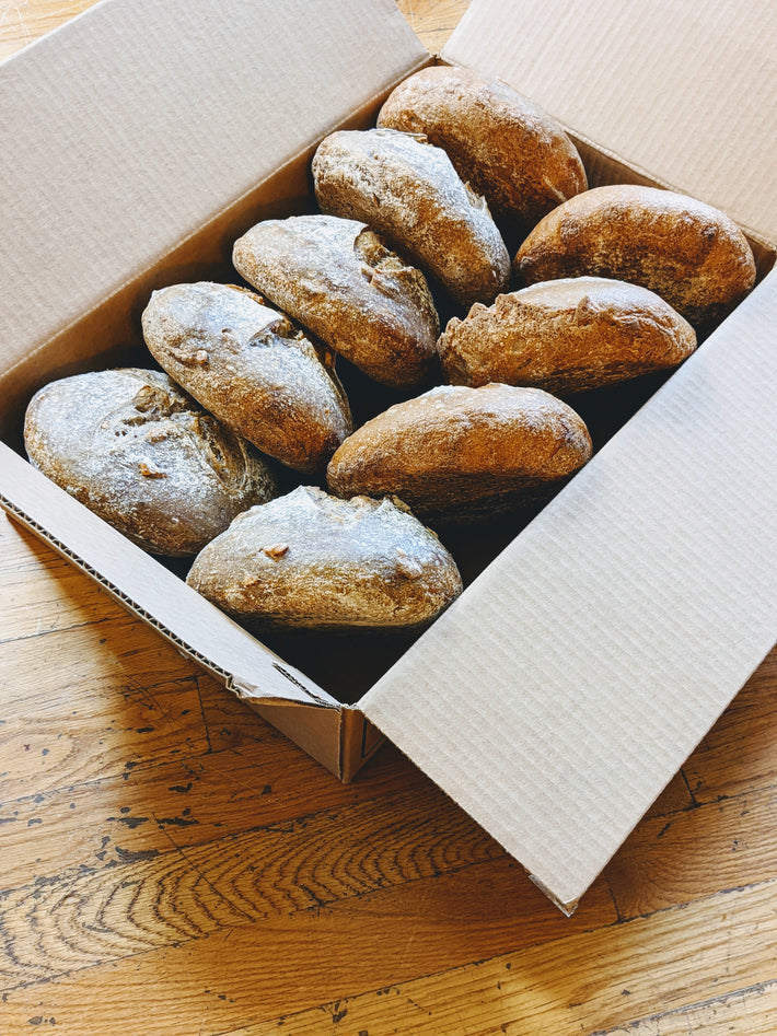 Cardboard box containing nine sourdough rolls dusted with flour, on a wooden floor.
