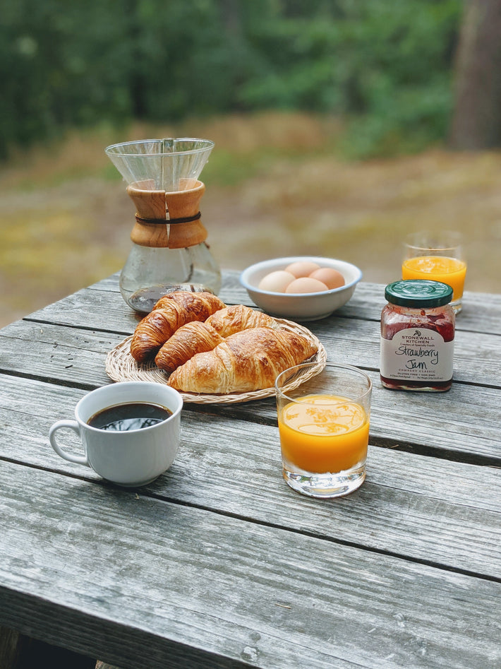 A rustic table set with croissants, coffee, orange juice, eggs, and strawberry jam.