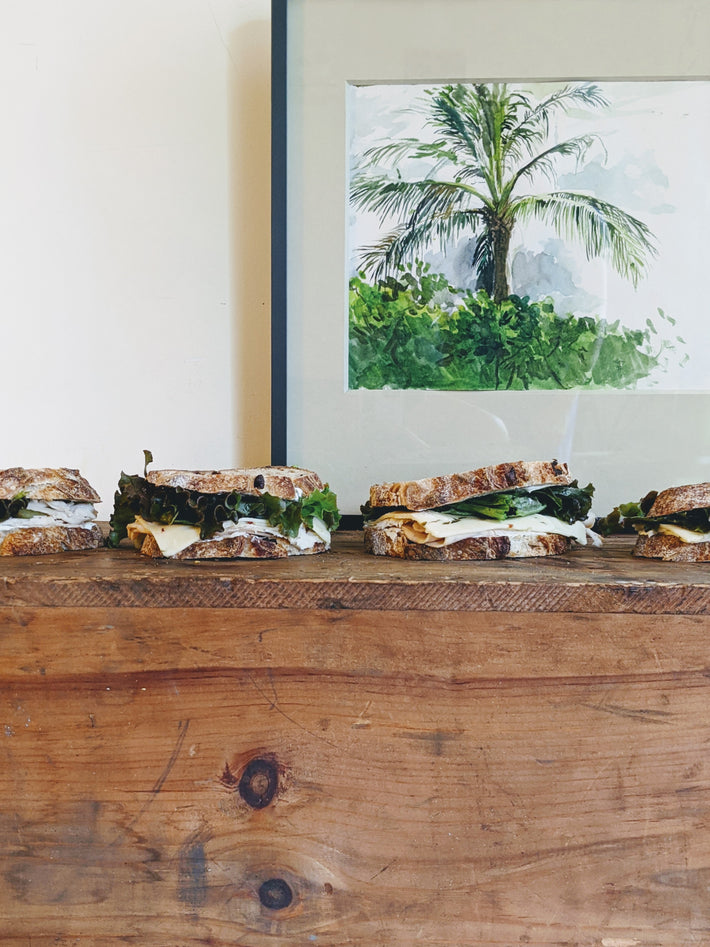 Four sandwiches on a wooden table with a palm tree painting in the background.