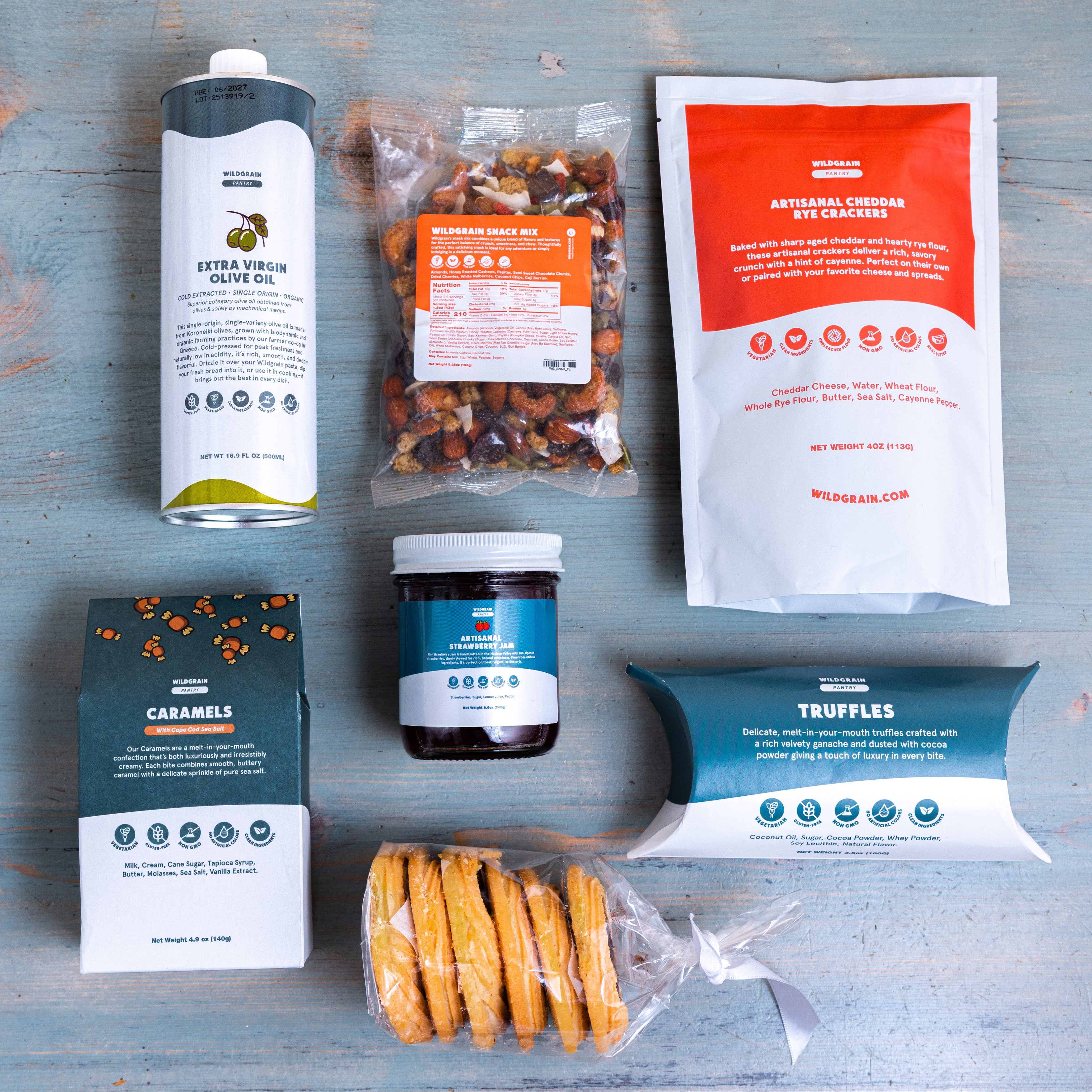 Wildgrain Treat Box items on wood: EXTRA VIRGIN OLIVE OIL CARAMELS TRUFFLES ARTISANAL CHEDDAR WILDGRAIN.COM