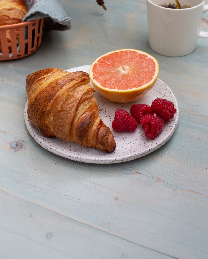 Croissant on a speckled plate with half grapefruit, raspberries, and a white coffee mug on a pale wooden table.