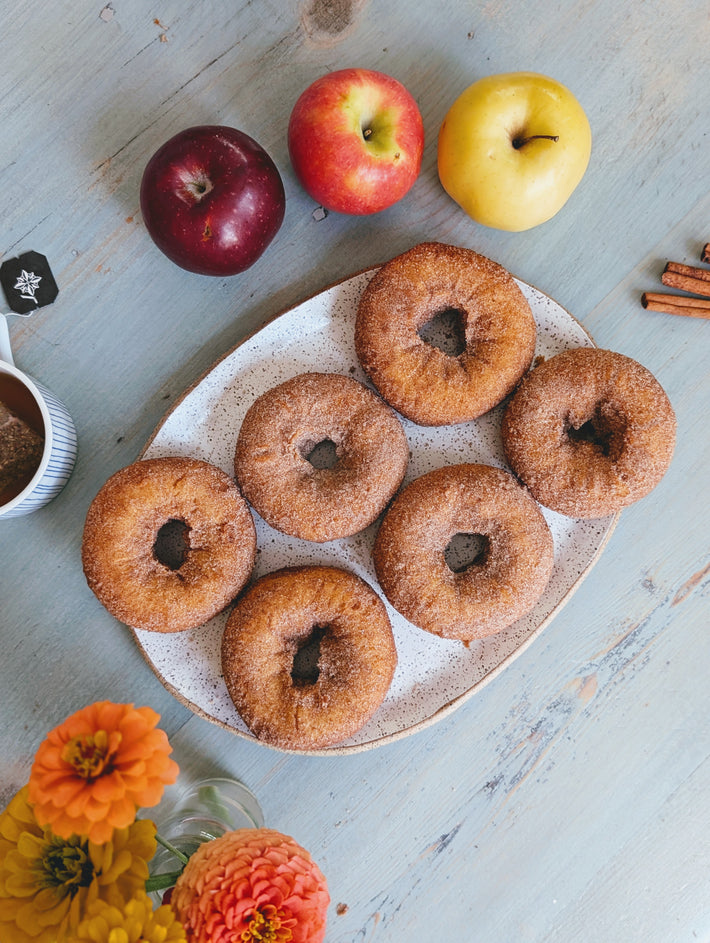 A plate of six cinnamon sugar donuts surrounded by apples, flowers, and a cup of coffee on a wooden table.