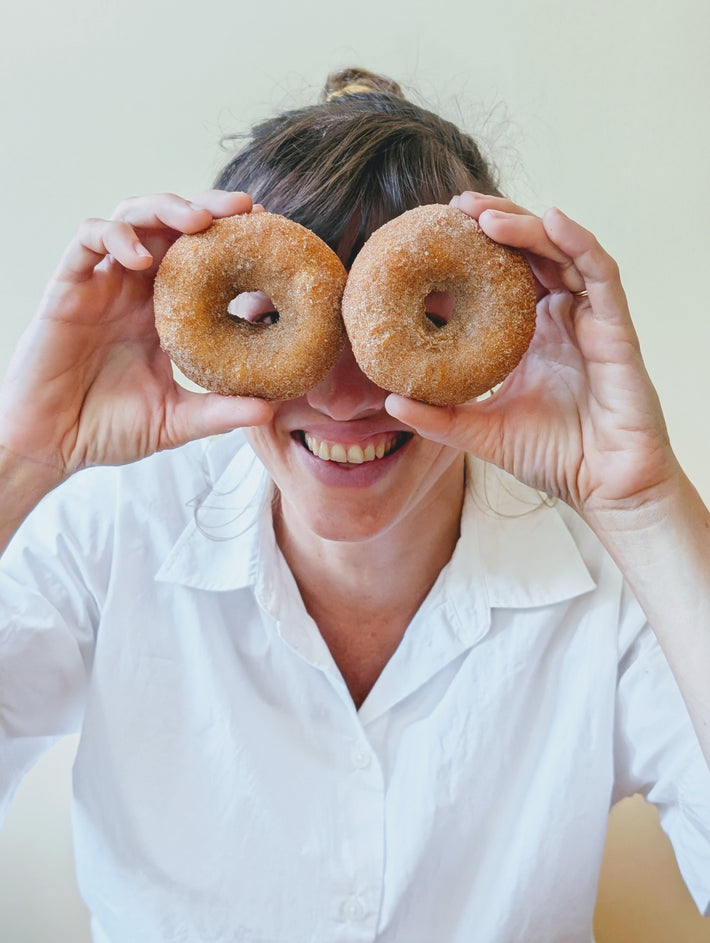 A person holds two Apple Cider Donuts in front of their face, smiling. They wear a white shirt.