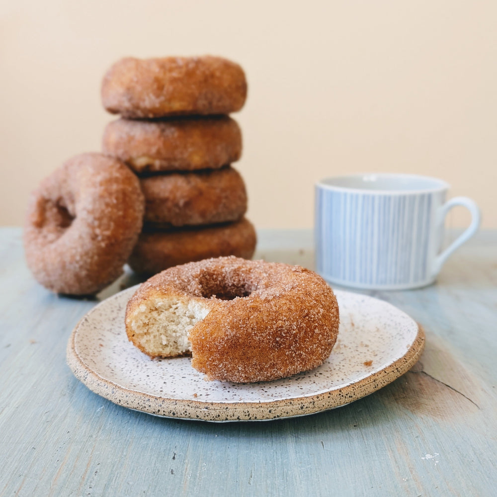 Close-up of cinnamon-sugar donut with a bite showcasing sugar coating on a ceramic plate, stacked donuts and a coffee cup on a pale blue wooden table