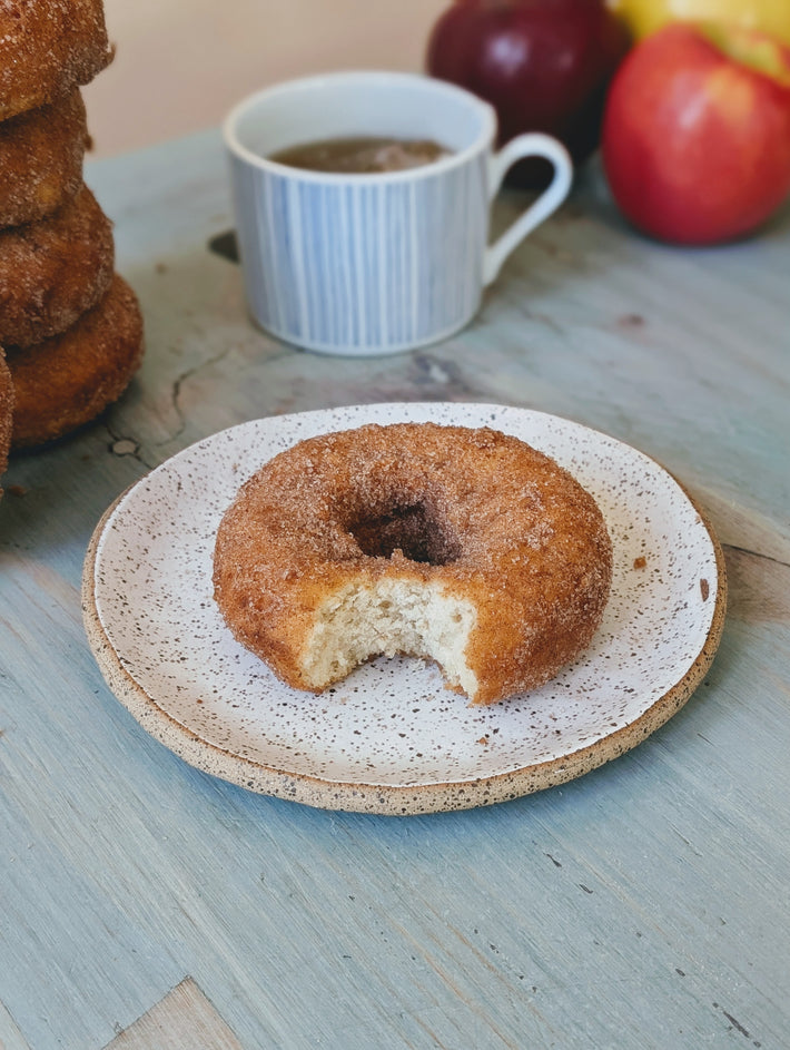 Cinnamon sugar donut with a bite on a speckled plate, coffee cup and apples in background.