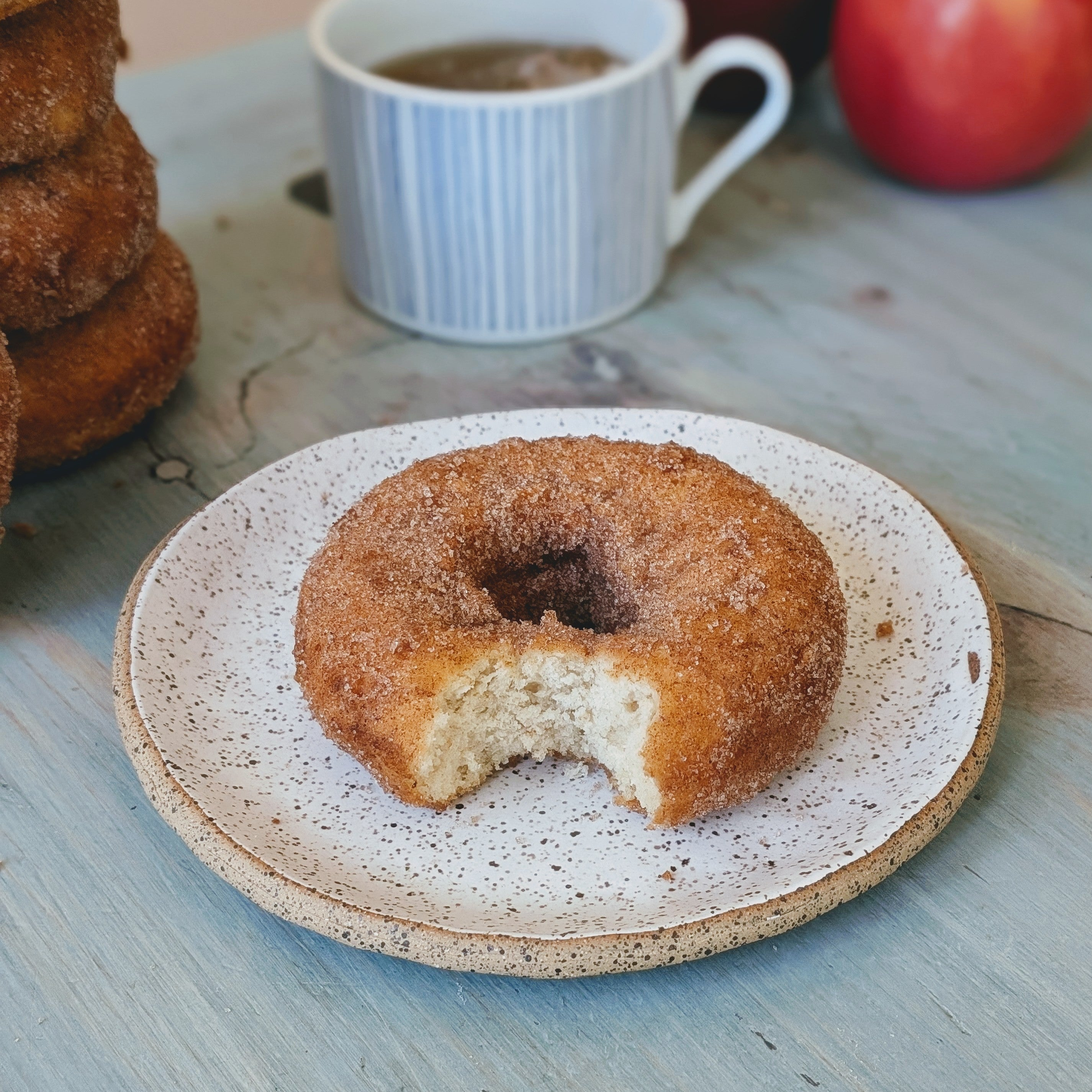 Single apple-cider donut with a bite, on a speckled plate with a coffee cup and apple in the background.