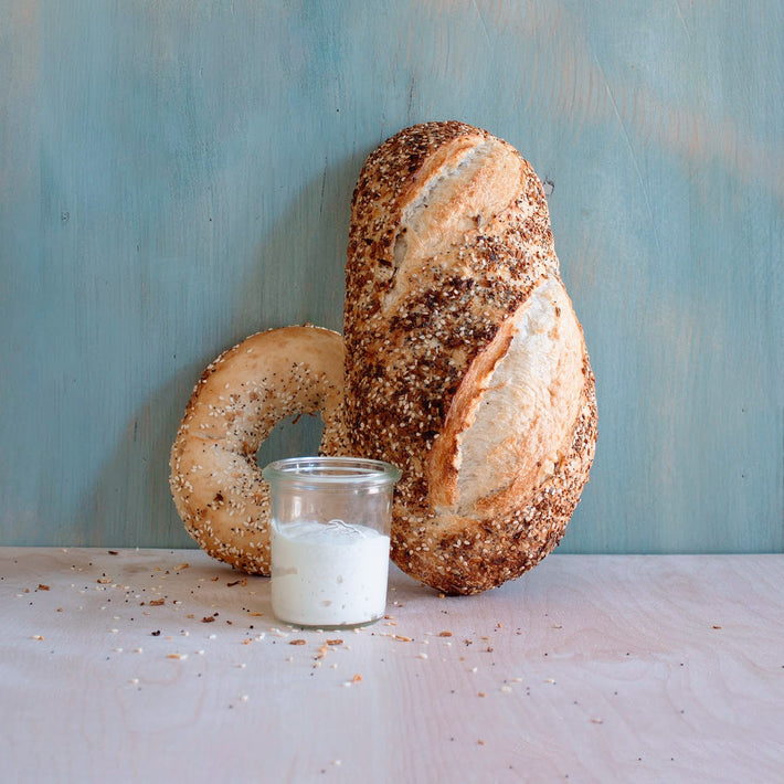 A loaf of seeded bread, a sesame bagel, and a small jar of white spread on a wooden surface with a blue background.