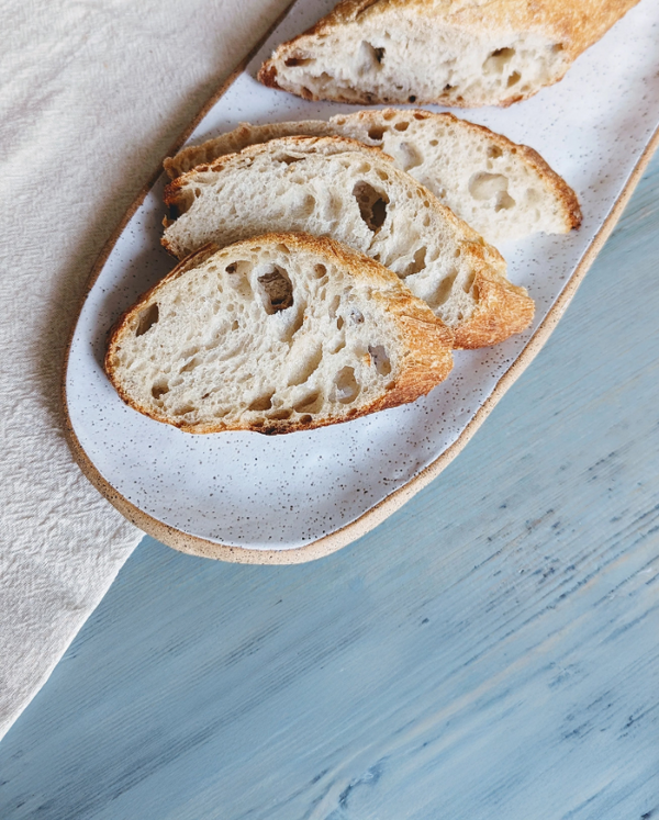 Four slices of crusty sourdough on a speckled oval plate beside a linen napkin on a light blue wood table.