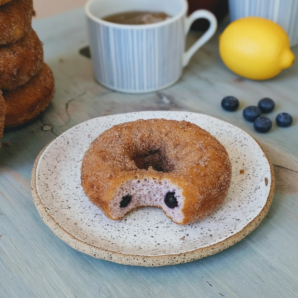 Cinnamon-sugar donut with a bite revealing blueberry-filled interior on a speckled ceramic plate, coffee mug, lemon and blueberries nearby.