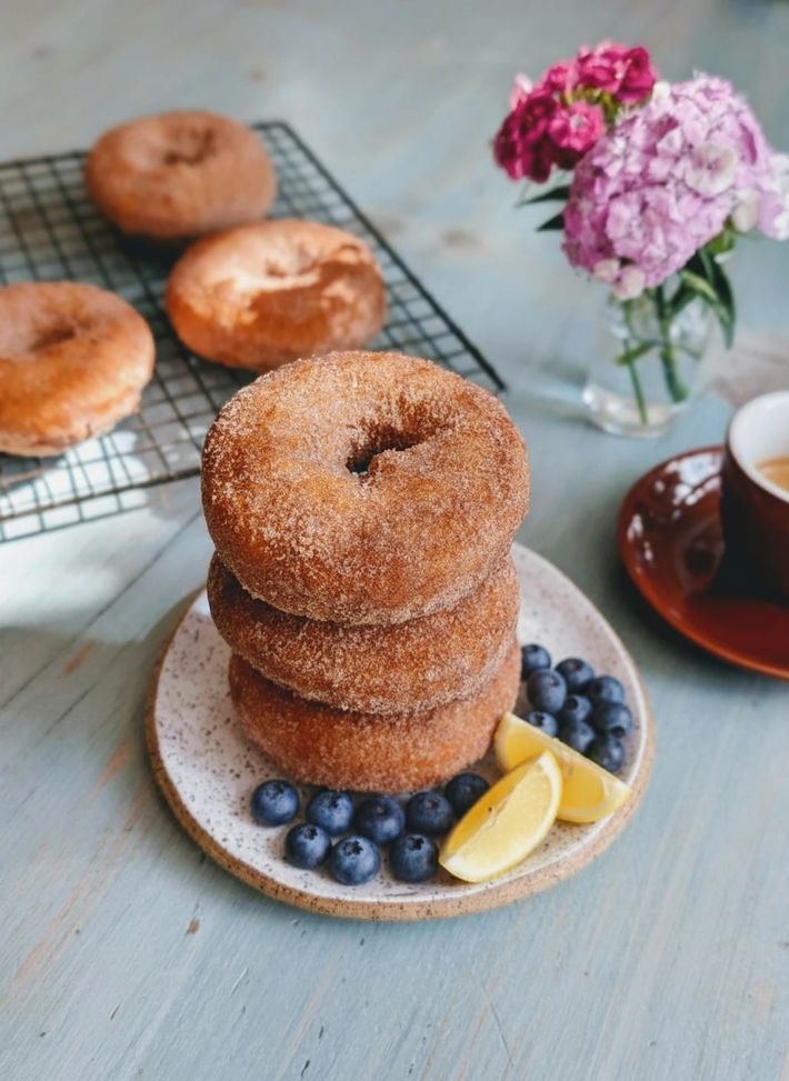Stacked cinnamon-sugar donuts on a plate with blueberries and lemon wedges, beside a coffee cup and vase of pink flowers.