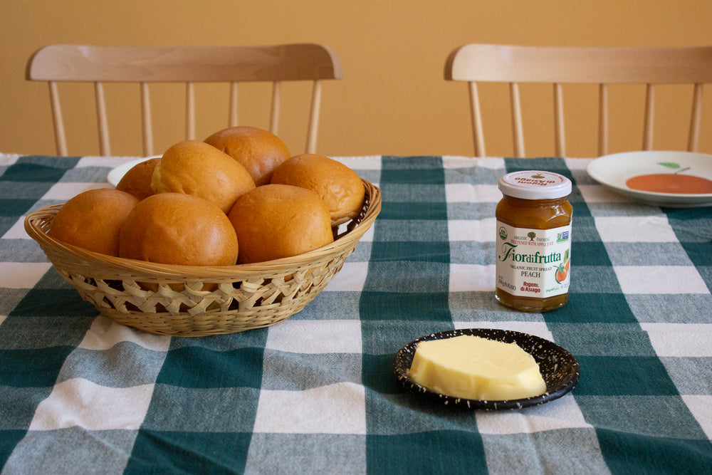 Basket of brioche rolls on checked tablecloth, jar labeled 'Fiordifrutta PEACH Rigoni di Asiago', and plate with butter.