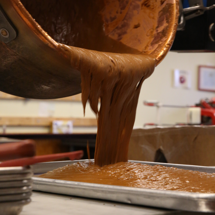Thick caramel being poured from a copper pot into a shallow rectangular baking sheet in a kitchen.