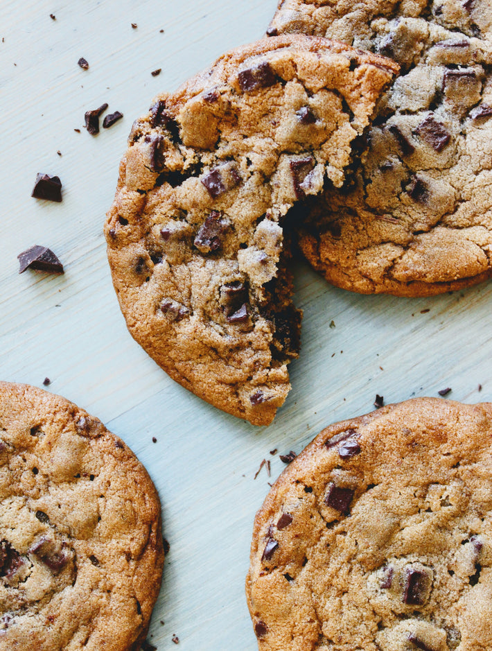 Two chocolate chip cookies on a pale blue surface, one broken to show gooey center and scattered chocolate chunks