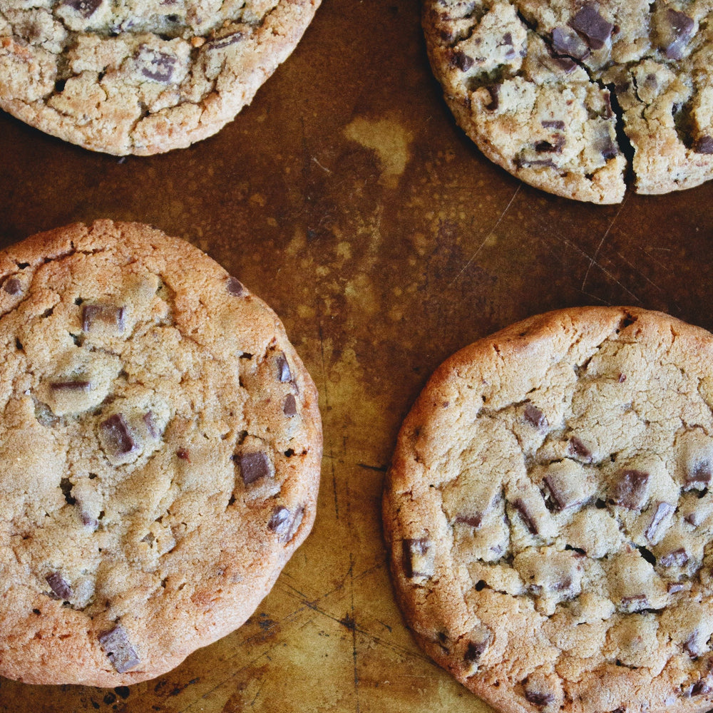 Four large chocolate-chunk cookies on a worn baking sheet, close-up showing texture and chocolate pieces