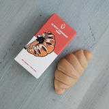 Angled top-down view of Le Petit Croissant Soap showing croissant-shaped tan soap and its illustrated box on light gray wood