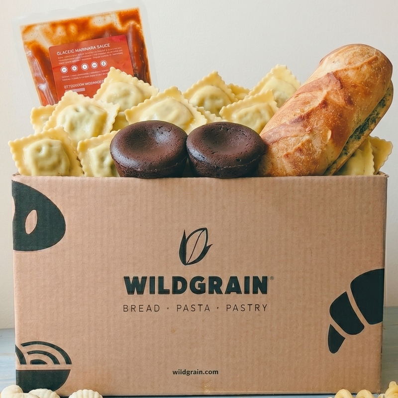Front view of Date Night Box with Wildgrain cardboard box filled with ravioli, marinara pouch, garlic butter baguette, and two chocolate lava cakes on a light background