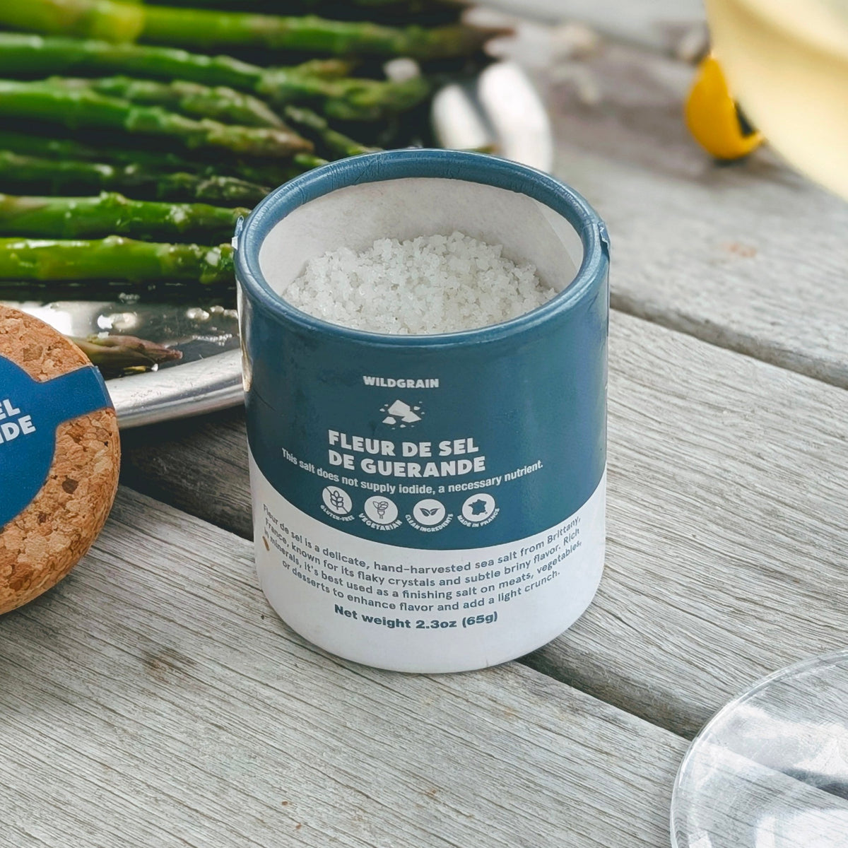 A cylindrical tin of Fleur de Sel de Guerande sea salt sits on a weathered gray wooden table, surrounded by fresh green asparagus, a glass of white wine, and a cork coaster.