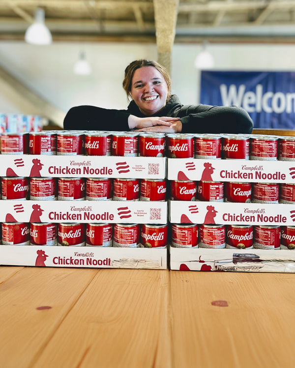 Smiling volunteer leaning on stacked Campbell's Chicken Noodle cases with a blurred 'Welcome' banner behind.