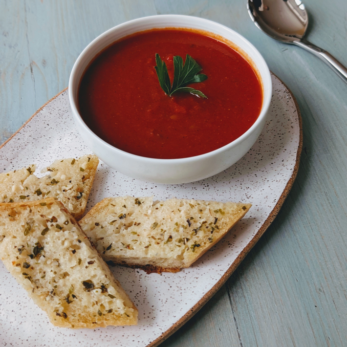 Angled close-up of Garlic Butter demi baguette slices showing herbed garlic butter topping on a speckled plate with a bowl of tomato soup.