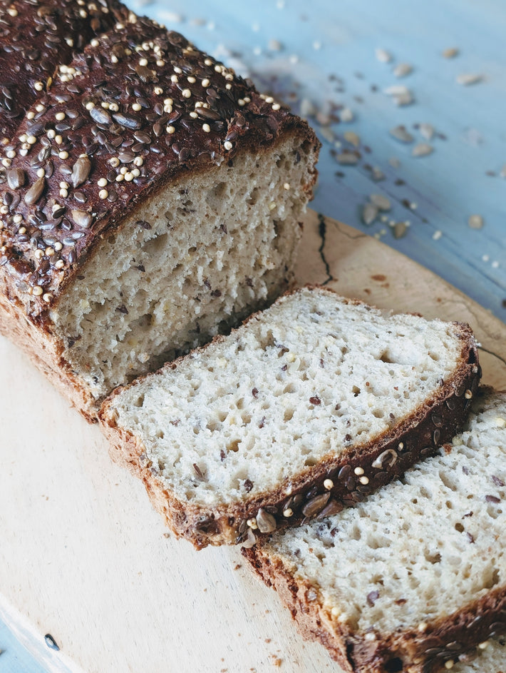 Gluten-Free Slow-Fermented Seeded Loaf, partially sliced on a wooden board, with crumbs scattered around.