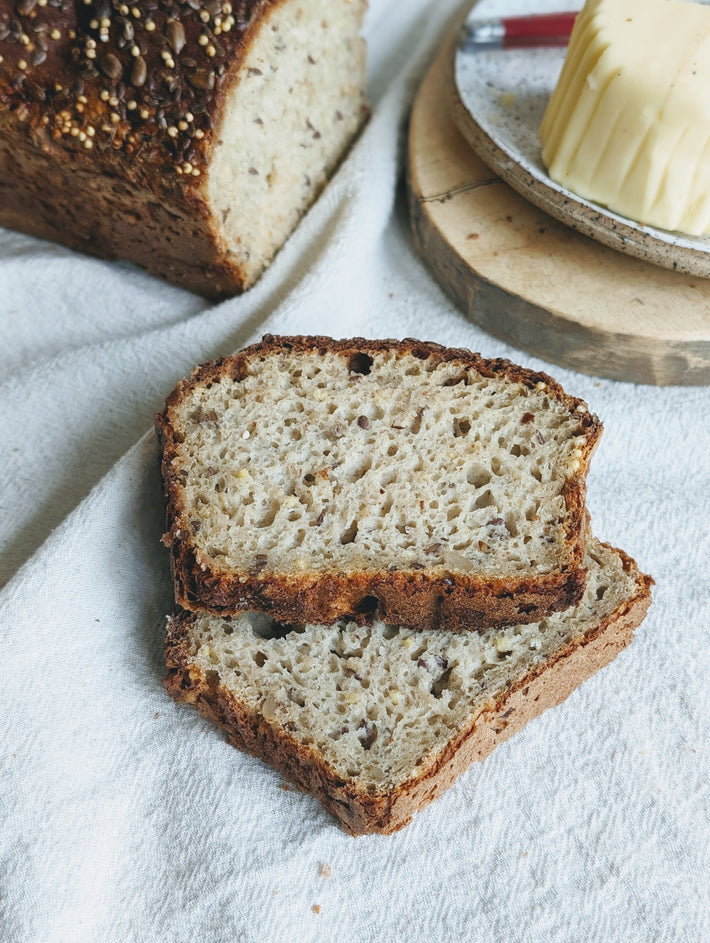 Gluten-Free Slow-Fermented Seeded Loaf sliced on a white cloth with a whole loaf and butter in the background.