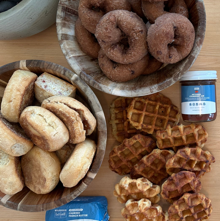 Top-down of Heat and Serve Catering Box: donuts, English muffins, maple waffles, jar of strawberry jam, and butter on wood