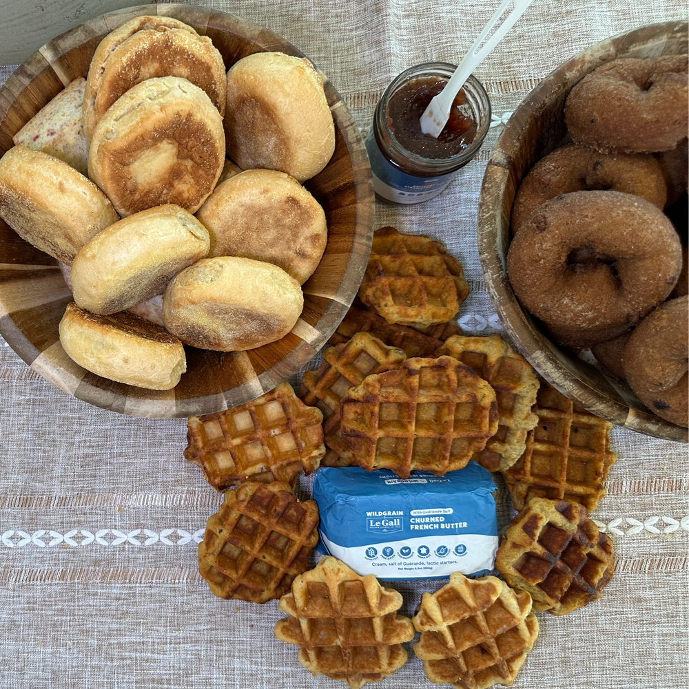 Assorted artisan donuts, bagels, English muffins, and mini waffles with butter and jam arranged on a table ready to heat and serve