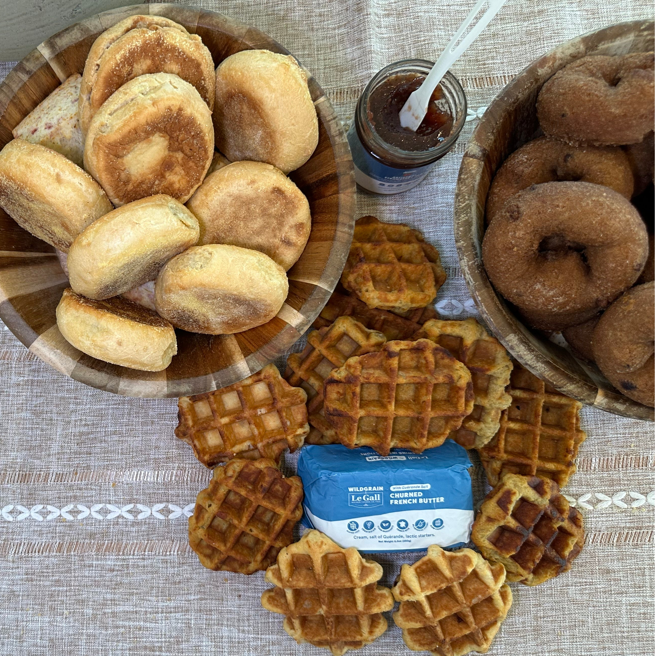 Assorted artisan donuts, bagels, English muffins, and mini waffles with butter and jam arranged on a table ready to heat and serve