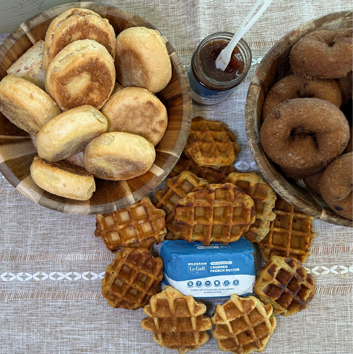 Assorted artisan donuts, bagels, English muffins, and mini waffles with butter and jam arranged on a table ready to heat and serve