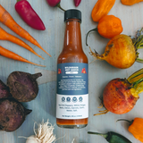 Front view of Wildgrain Hot Sauce bottle (Red, 5 fl oz) showcasing the label and sauce color with assorted fresh vegetables on a light wood background