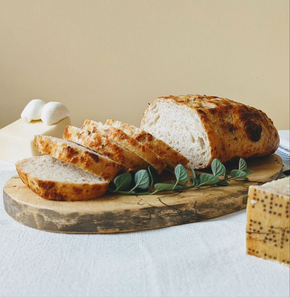 Sliced rustic sourdough loaf on a wooden board with fresh herbs, two mozzarella balls and a wedge of peppered cheese