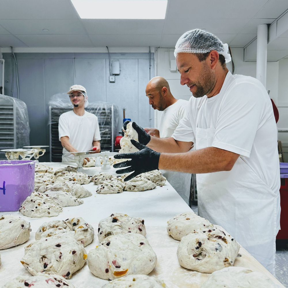 Three bakers in hairnets shaping loaves of fruit-and-nut dough on a long table in a commercial bakery.