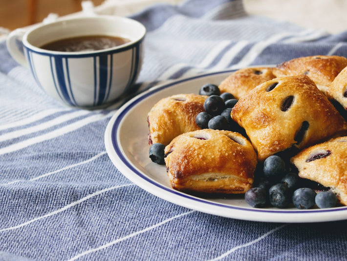 A plate of blueberry pie bites with fresh blueberries and a cup of coffee on a striped cloth