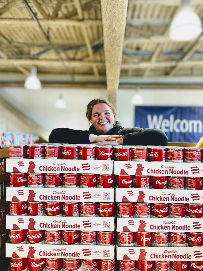 Smiling volunteer leaning on stacked Campbell's Chicken Noodle soup cases with Welcome banner behind.