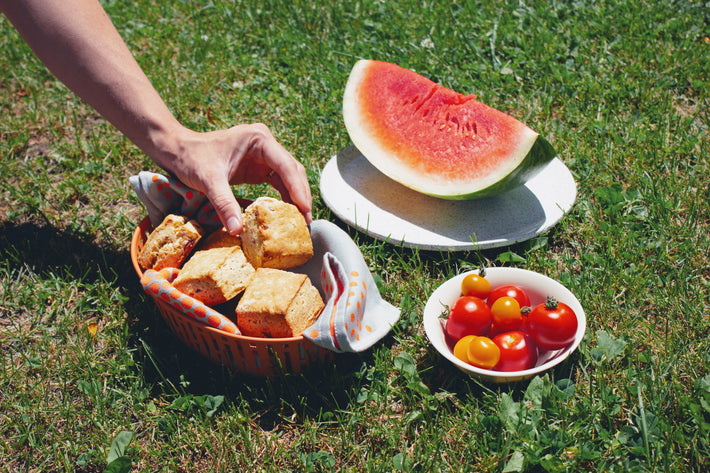 A hand reaching for biscuits in a basket next to a watermelon and cherry tomatoes on a plate.