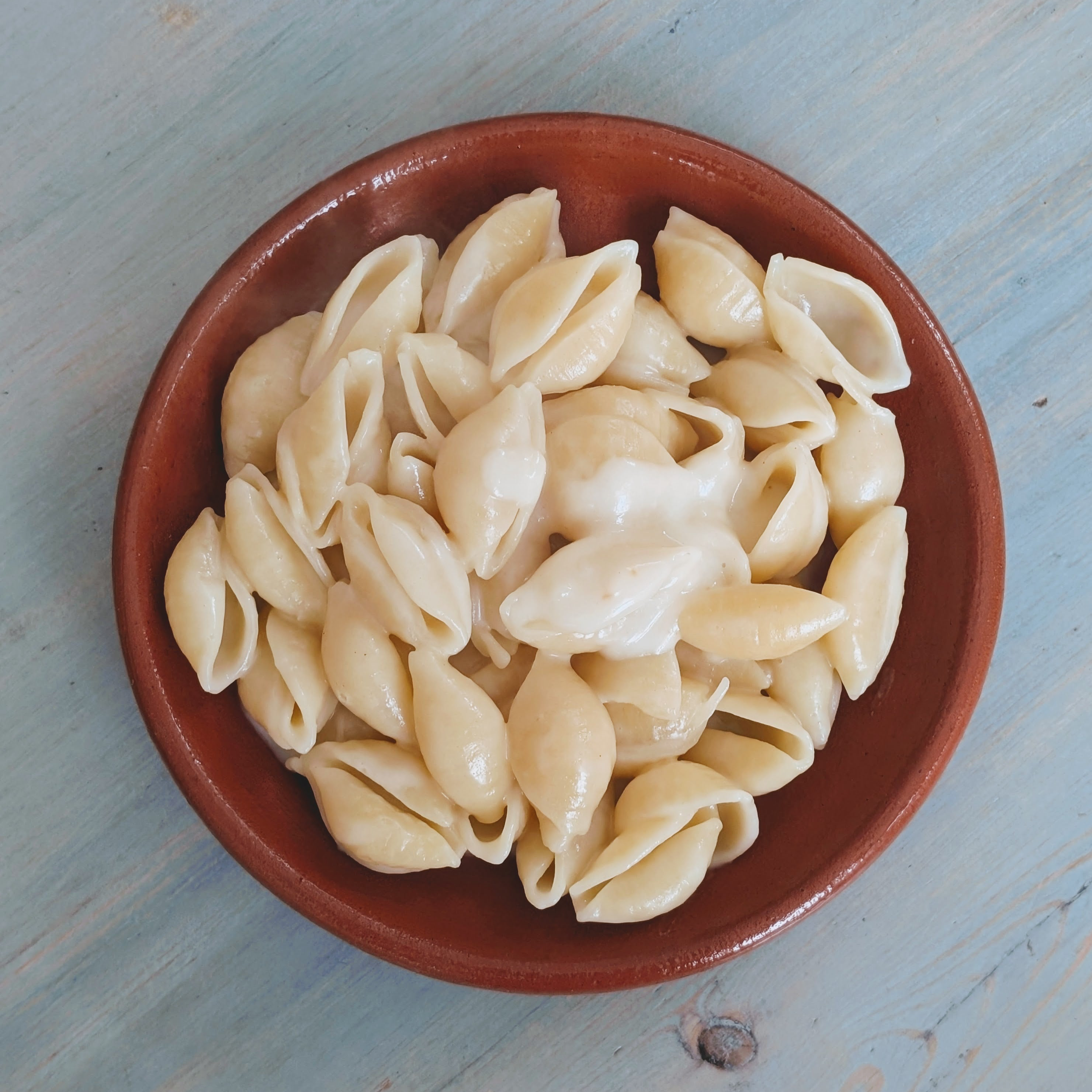 Top-down view of gluten-free shell pasta with creamy three-cheese sauce in a brown bowl on a pale blue wooden table
