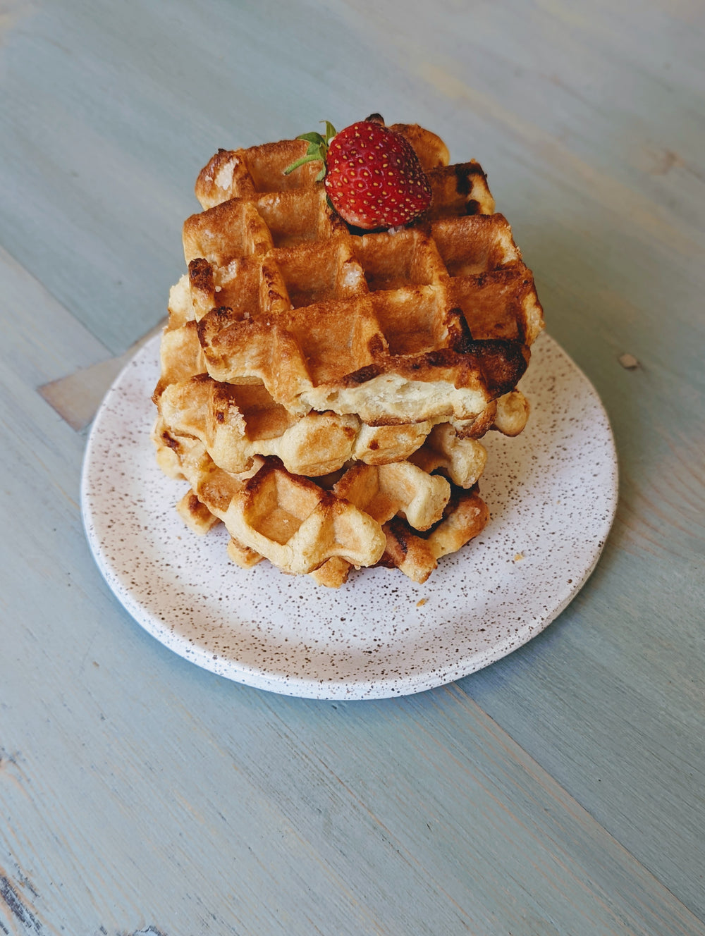 Three-quarter view of stacked maple Belgian waffles topped with a strawberry on a speckled plate on a blue wooden table