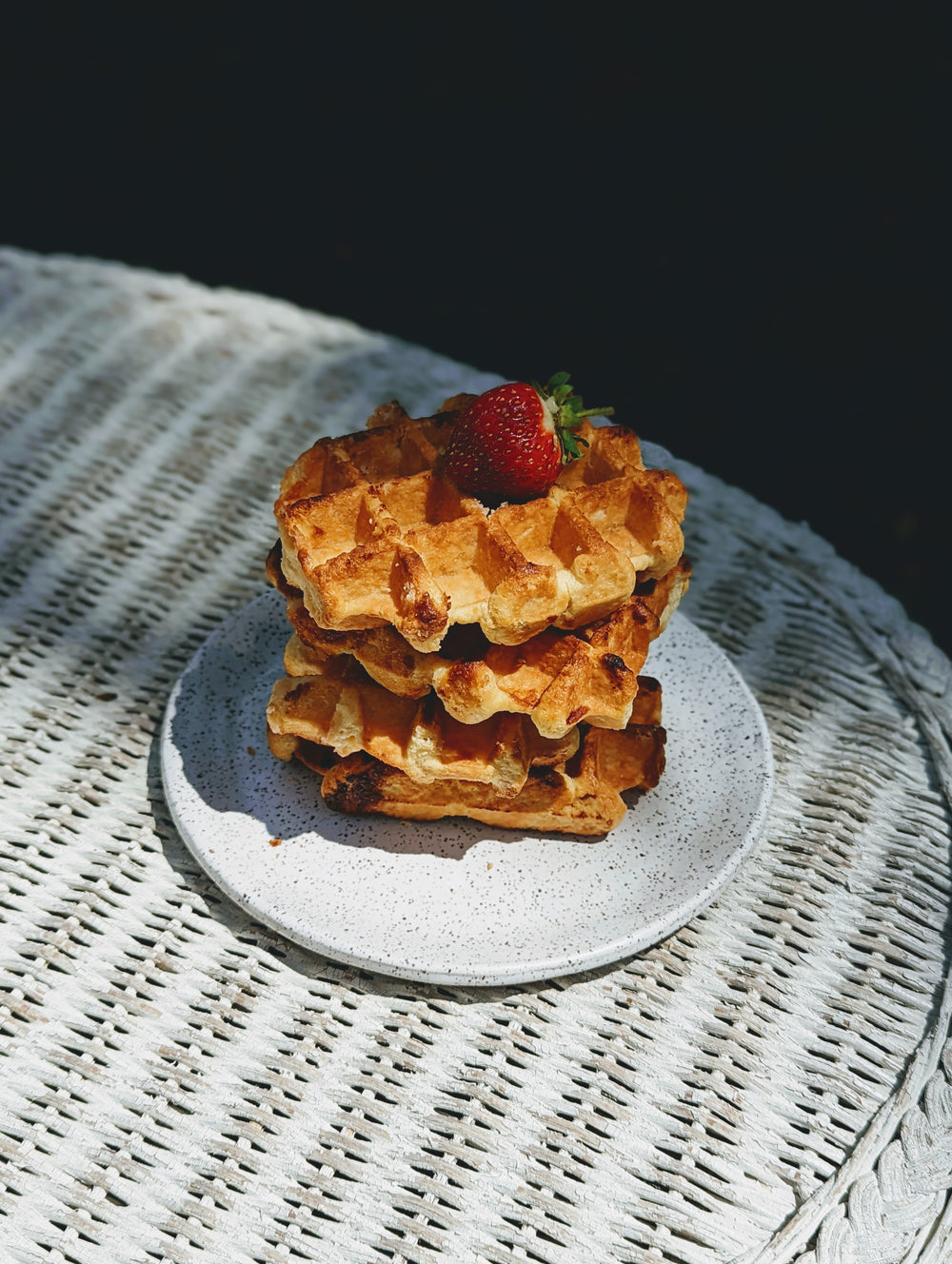 Angled overhead view of three Belgian maple waffles stacked on a speckled plate with a strawberry on a white wicker table