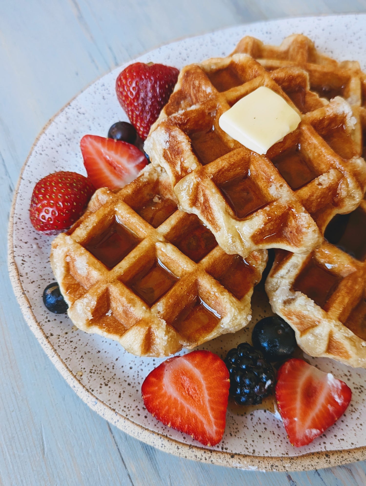 Top-down view of Belgian maple waffles stacked with melting butter and syrup, served with mixed berries on a speckled plate
