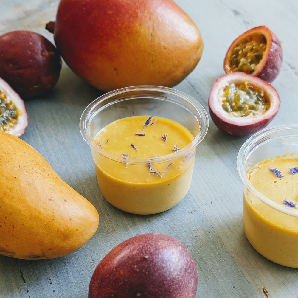 Two cups of mango–passionfruit mousse garnished with purple flower petals, surrounded by mangos and halved passionfruit on a wooden board.