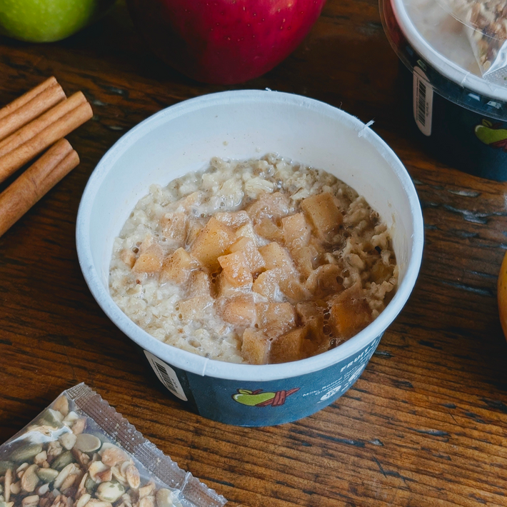 Top-down view of Fruits & Crunch Oats Apple & Cinnamon cup showcasing diced apples and creamy overnight oats on a wooden table with cinnamon sticks and a granola packet nearby