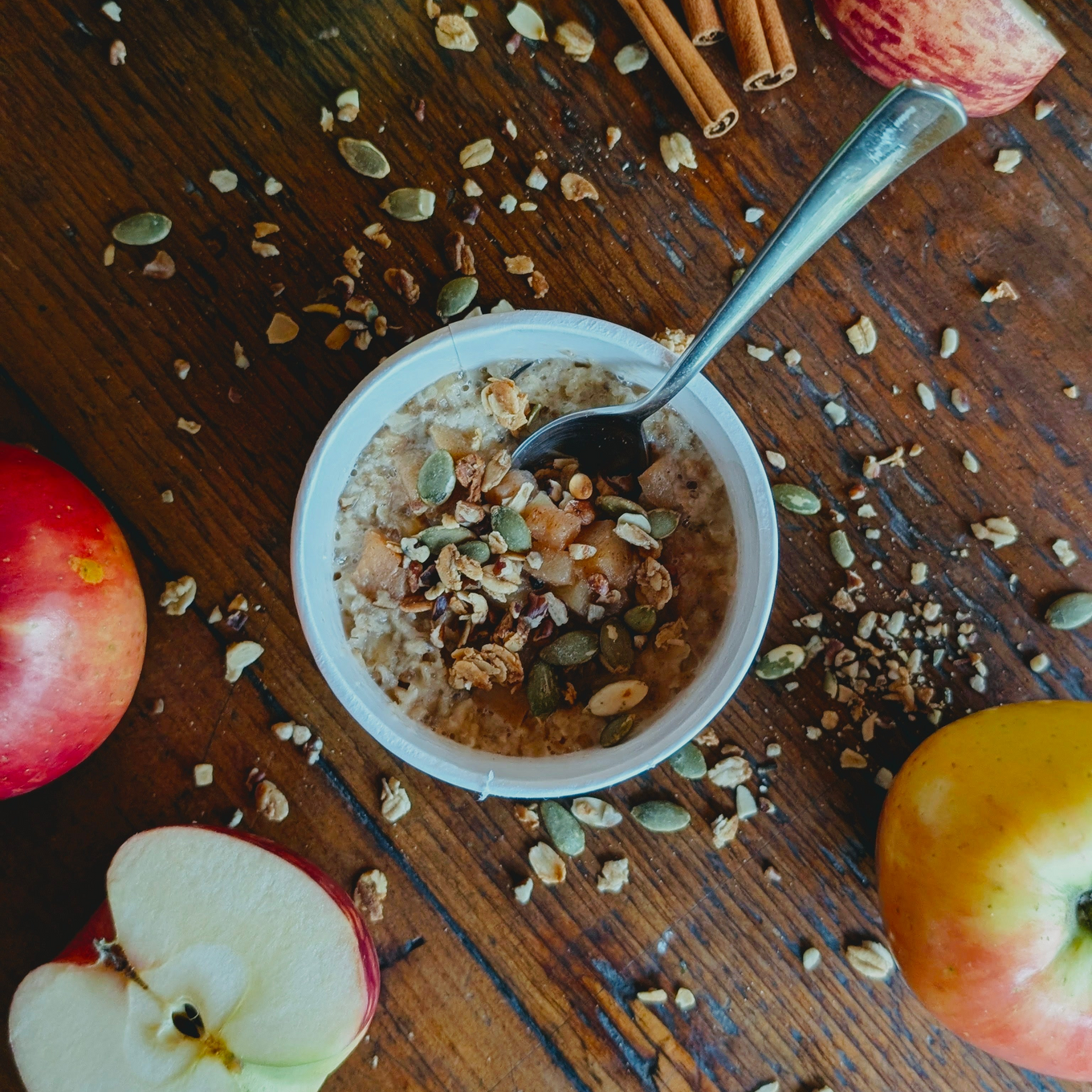 Top view of Fruits Crunch Oats Apple and Cinnamon cup showing spoon, granola, pumpkin seeds and apple slices on wooden table